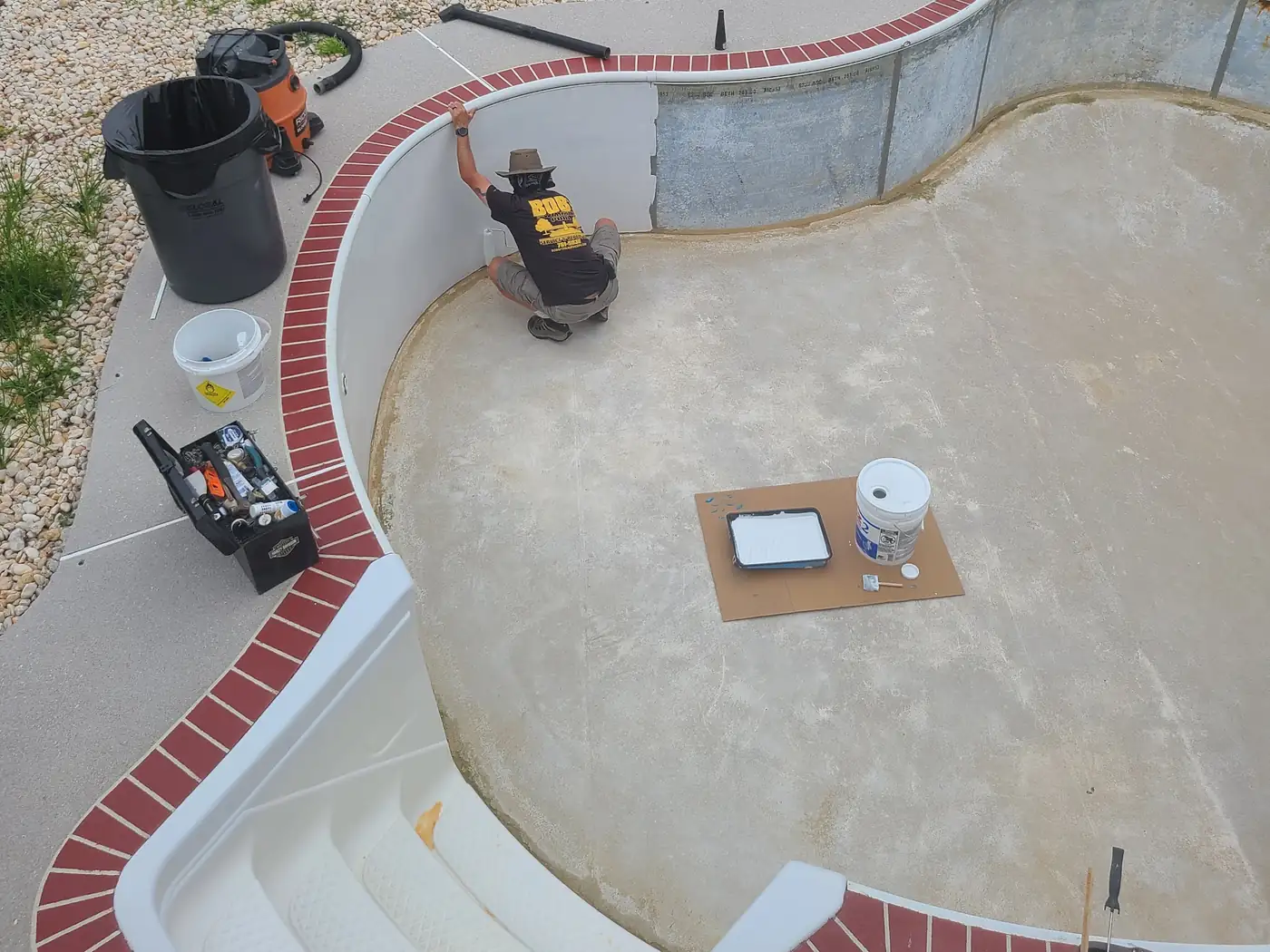 A Bob's crew member in a black and gold shirt priming the steel walls of an empty kidney pool, bare panels beside painted ones